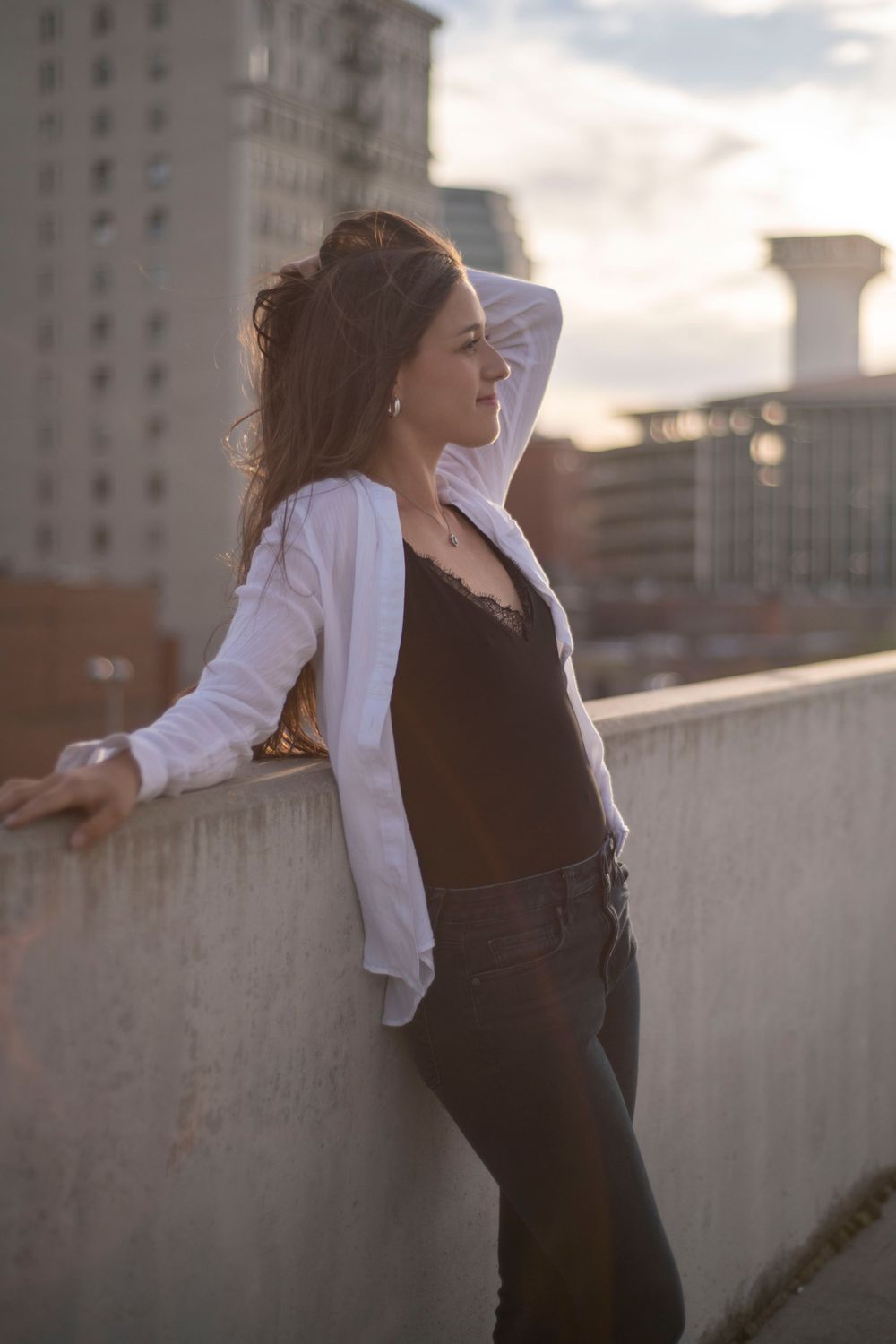Young woman in white cardigan and black top posing on rooftop with city buildings and cloudy sky.