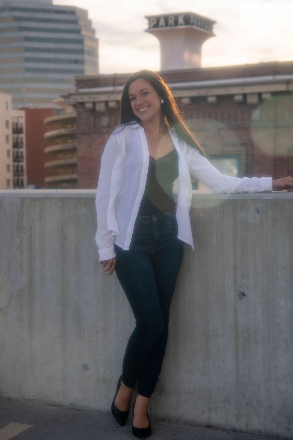 Woman in white shirt and black jeans leaning on rooftop ledge with urban Park building at sunset.