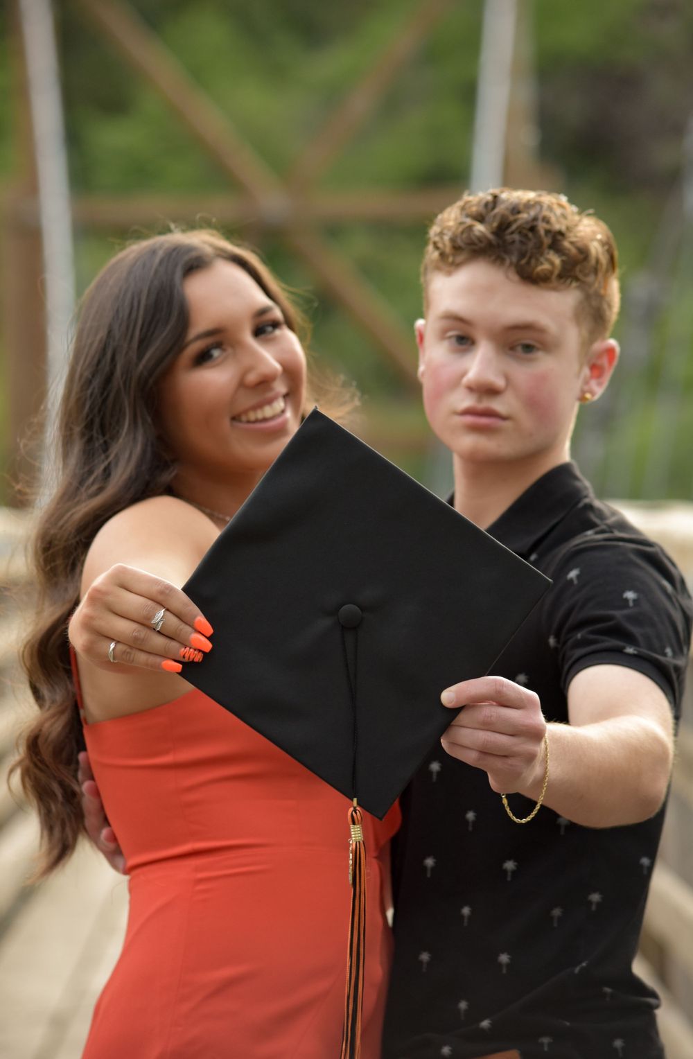 Young couple holds graduation cap toward camera on wooden bridge outdoors.