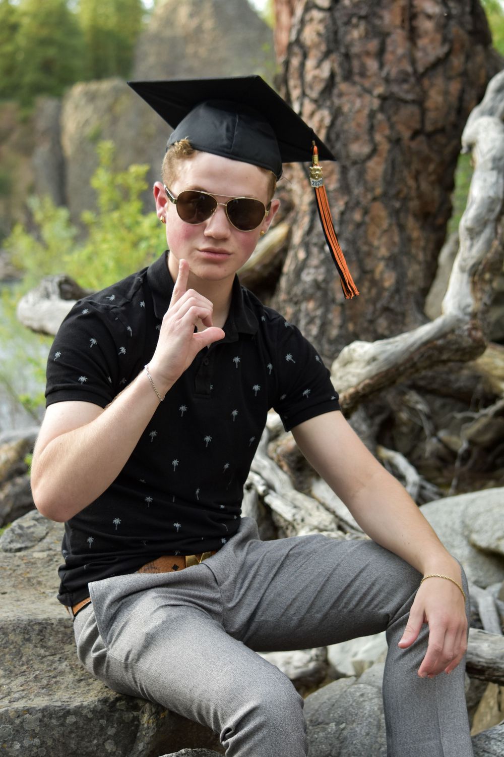 Young male graduate in black polo and graduation cap poses thoughtfully outdoors on rocks.