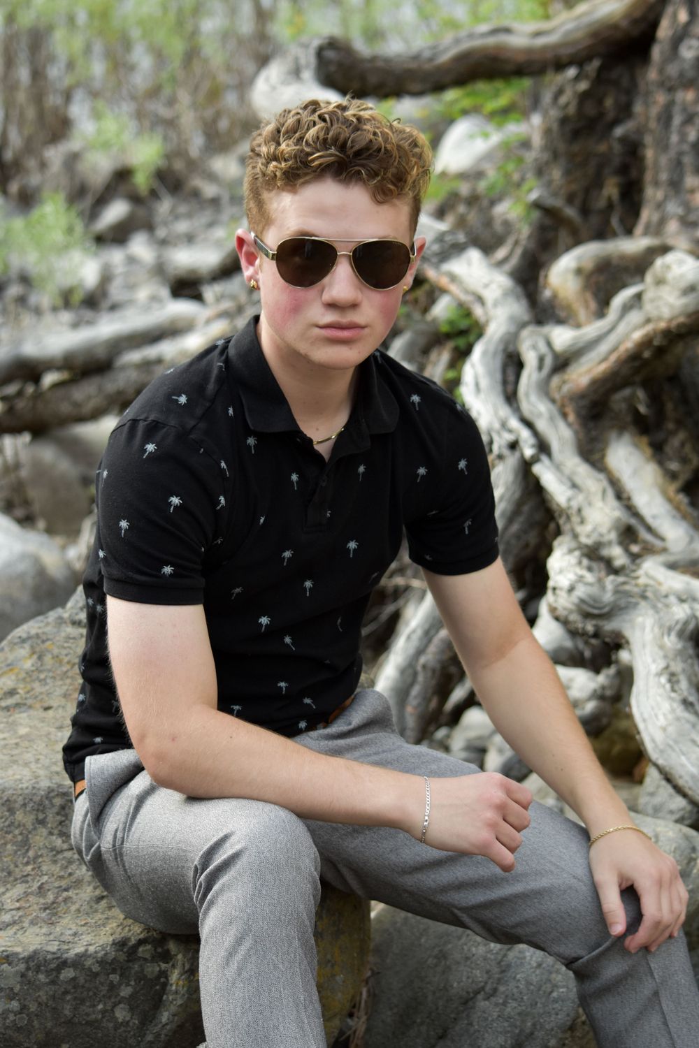 Young man with curly red hair wearing sunglasses and black polo shirt sitting on rocks outdoors.