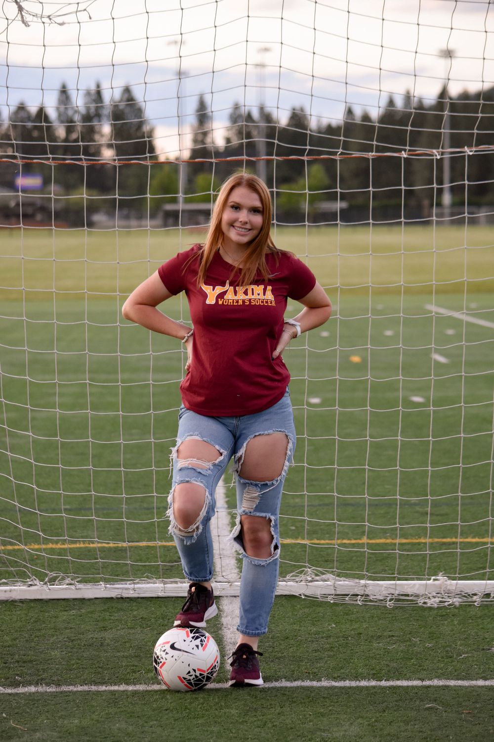 Young woman in Yakima soccer shirt leans on soccer ball at a goal net on a green turf field.