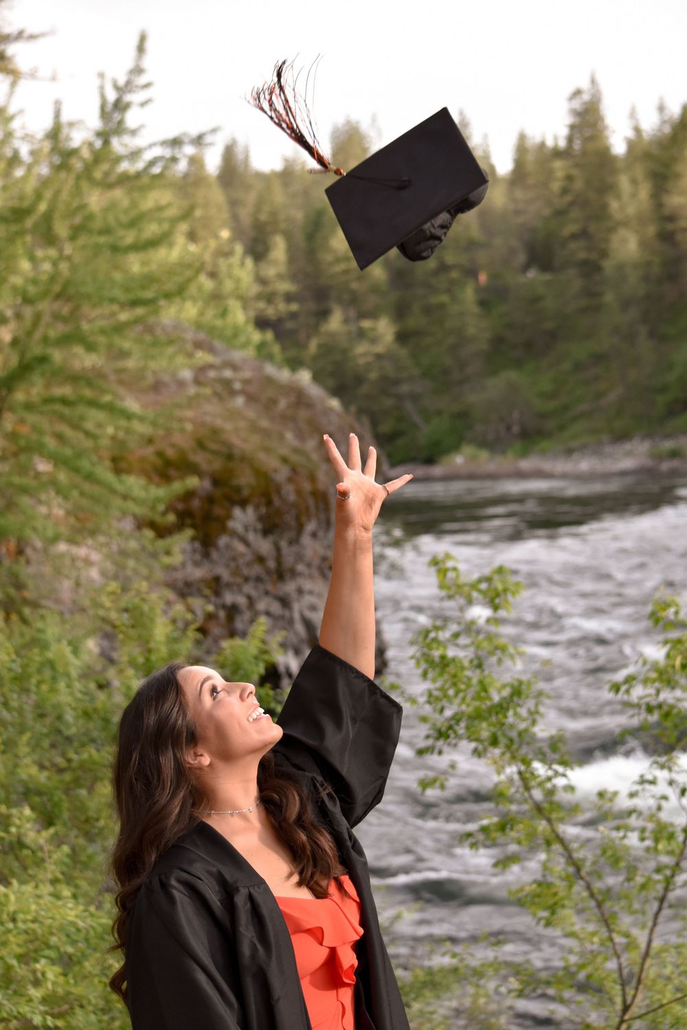 Smiling graduate in black gown tosses mortarboard cap into the air near a scenic river backdrop.