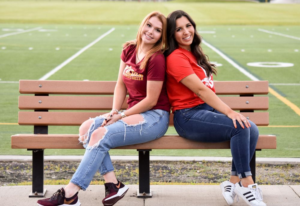 Two smiling young women sit back-to-back on bench near football field wearing school spirit shirts.