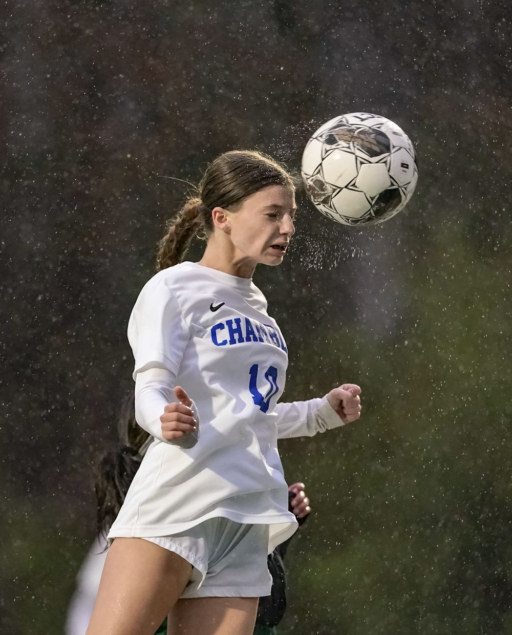 Soccer player heads the ball during rainy match wearing white uniform.