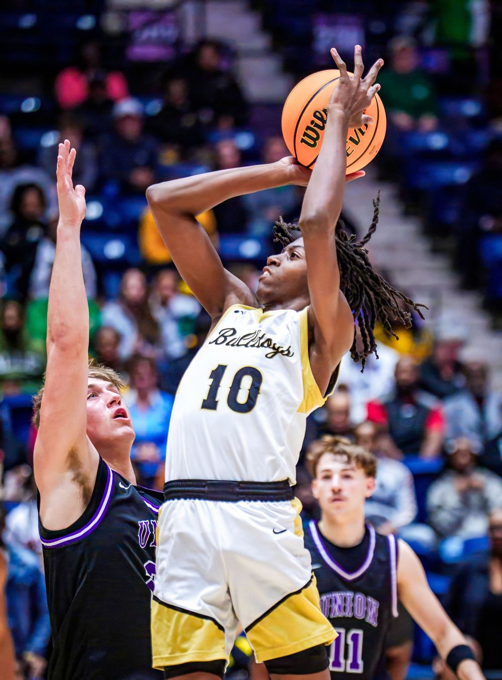 High school basketball player in yellow uniform takes a jump shot against defenders in purple jerseys during game action.
