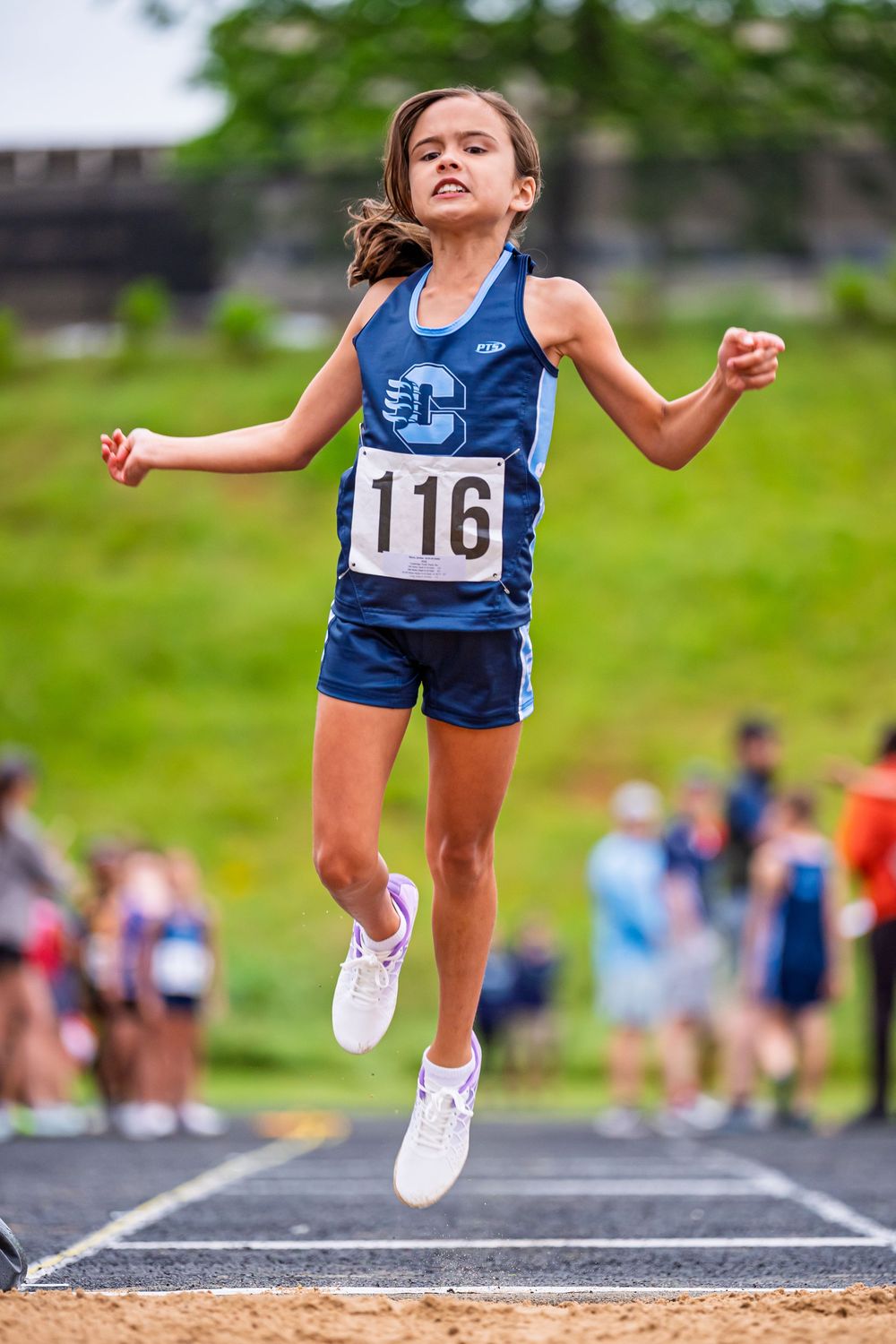 Young athlete in blue uniform competing in long jump at track and field event, leaping into sand pit with bib number 116.