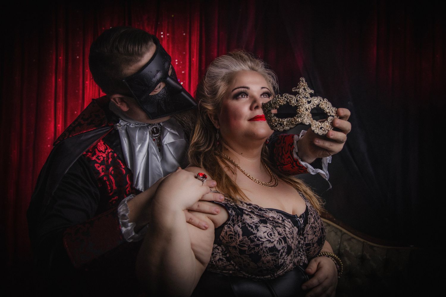 Dramatic gothic photoshoot with masked figure and decorative candelabra against red curtain backdrop.