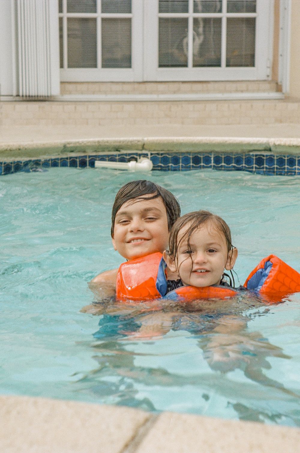 Two children wearing orange floaties swim together in a pool with blue tile edging.