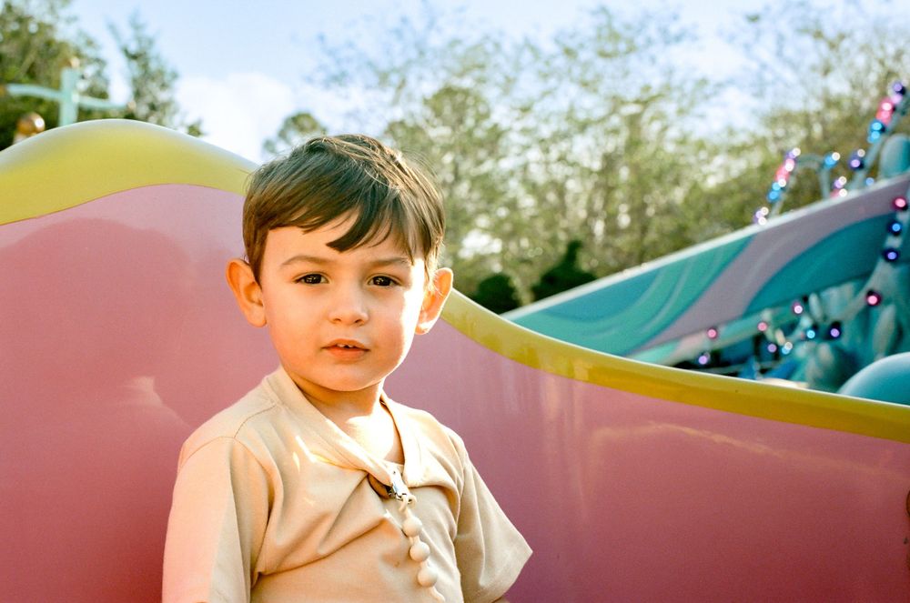 Young child in light colored shirt stands before a colorful playground structure on a sunny day.