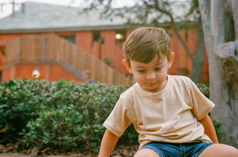 Young child in light colored t-shirt sitting outdoors with brick building and bushes in background.