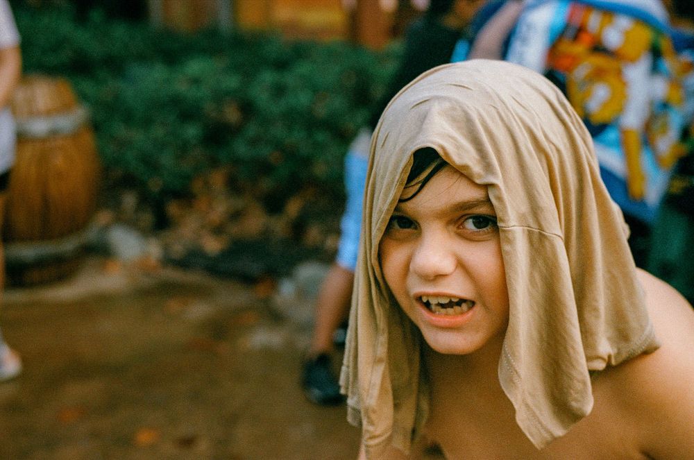 A person wearing a beige cloth draped over their head smiles playfully outdoors.