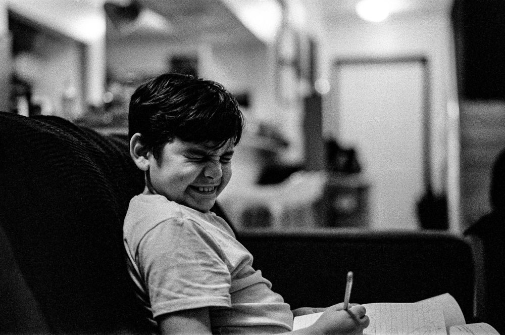 Black and white photo of a young person in a t-shirt writing or studying while seated at a desk.