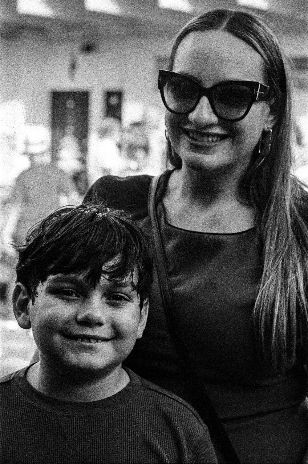 A black and white portrait of a young boy and woman smiling together, with the woman wearing dark sunglasses.