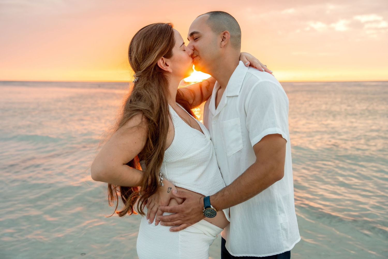 Happy couple sharing a kiss on the beach at sunset, woman in white dress with ocean backdrop.