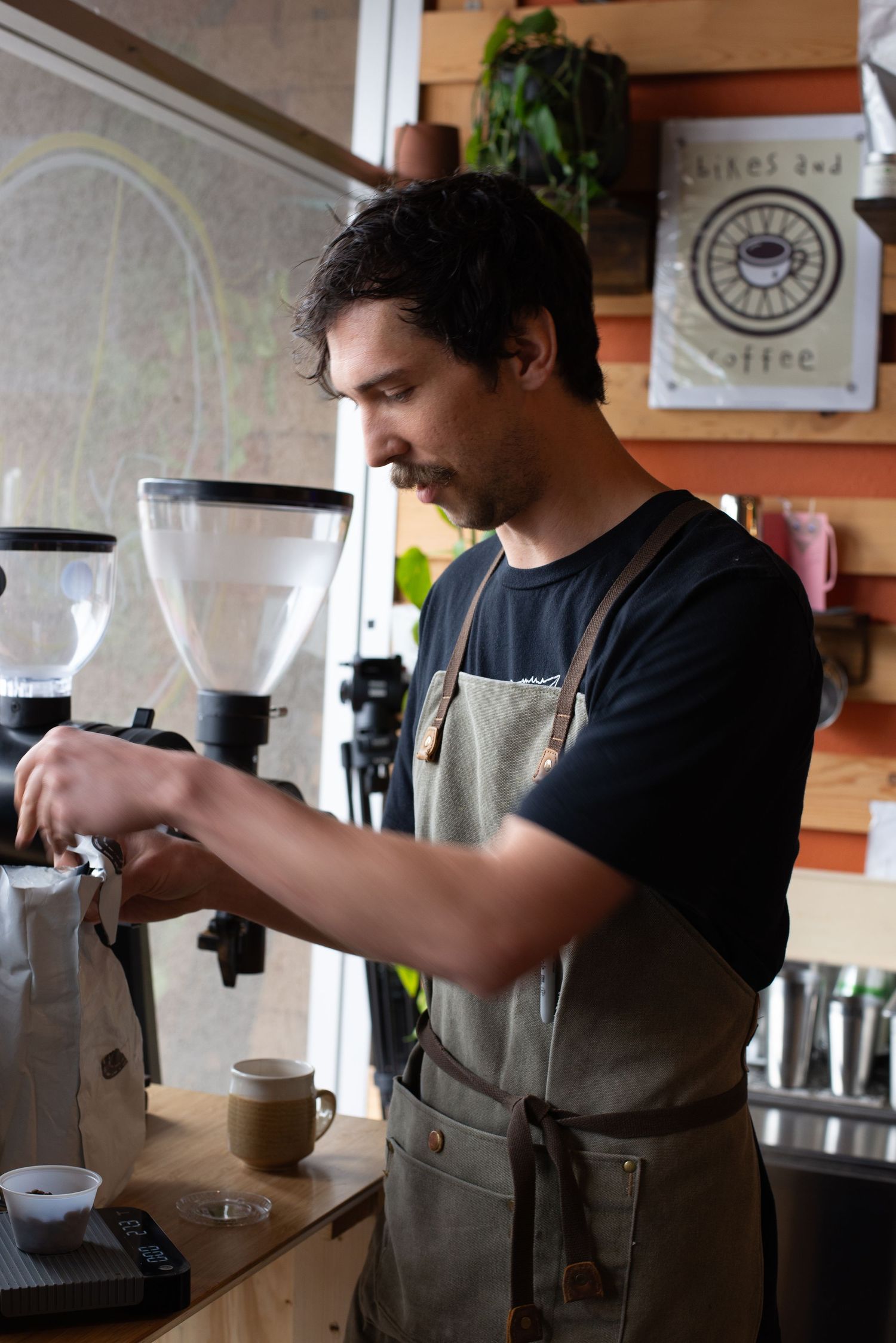 Male barista in a gray apron operating a coffee grinder at a specialty coffee shop counter.