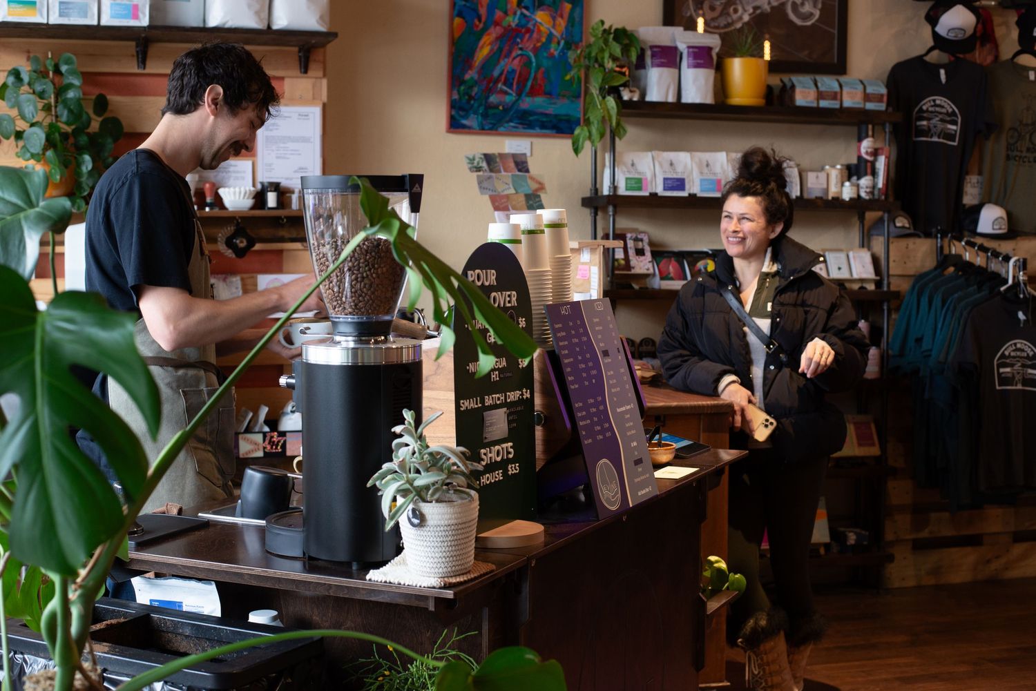 Two people sharing a cheerful conversation inside a cozy plant-filled coffee and retail shop.