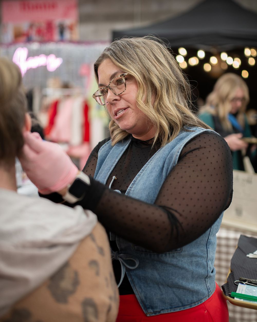 Woman with glasses and blonde hair wearing denim vest over black mesh top with red pants at an indoor market event.