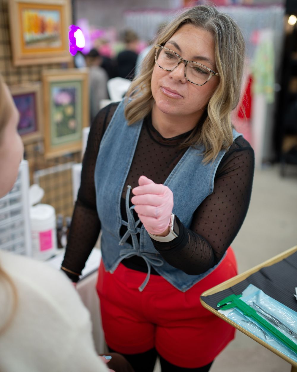 Female piercer wearing glasses and pink gloves preparing piercing tools at a decorated market booth.