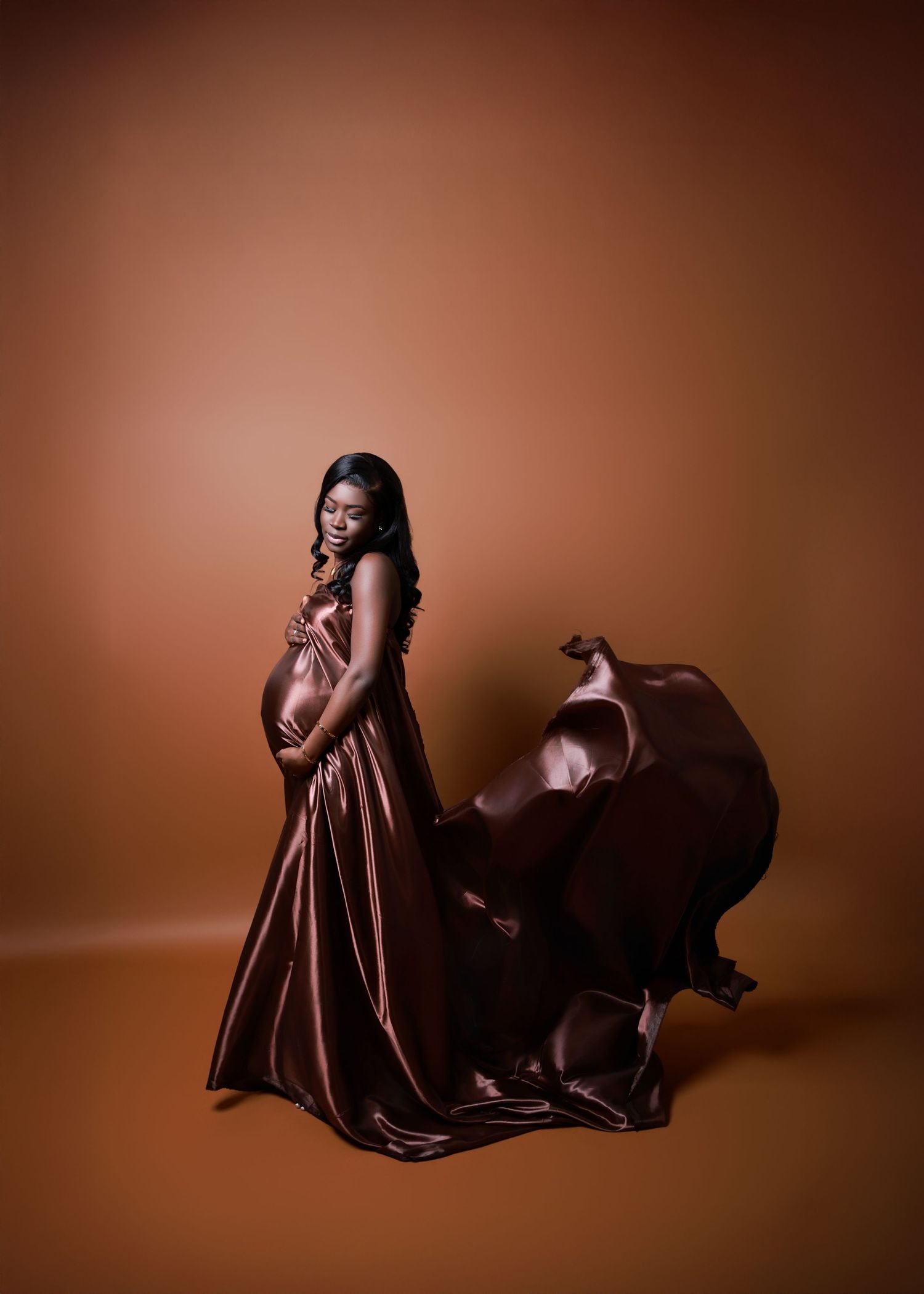 Artistic maternity photoshoot in flowing brown dress against warm backdrop.