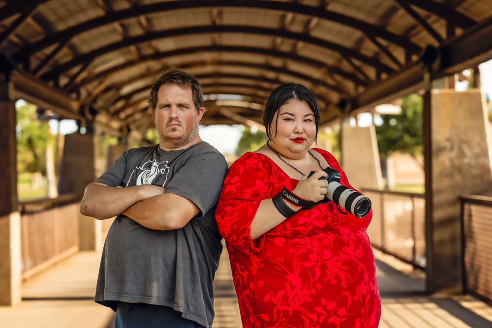 Two photographers pose back-to-back in a covered walkway, one holding a professional camera in a red dress.