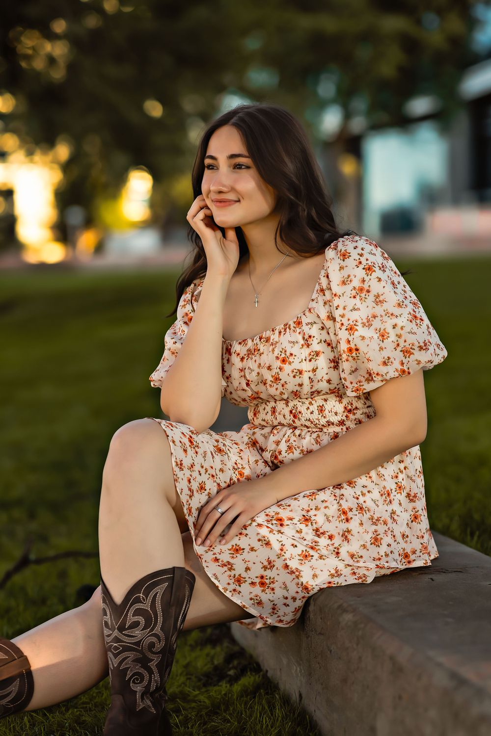 Woman in floral dress and cowboy boots poses on bench in sunset-lit park.