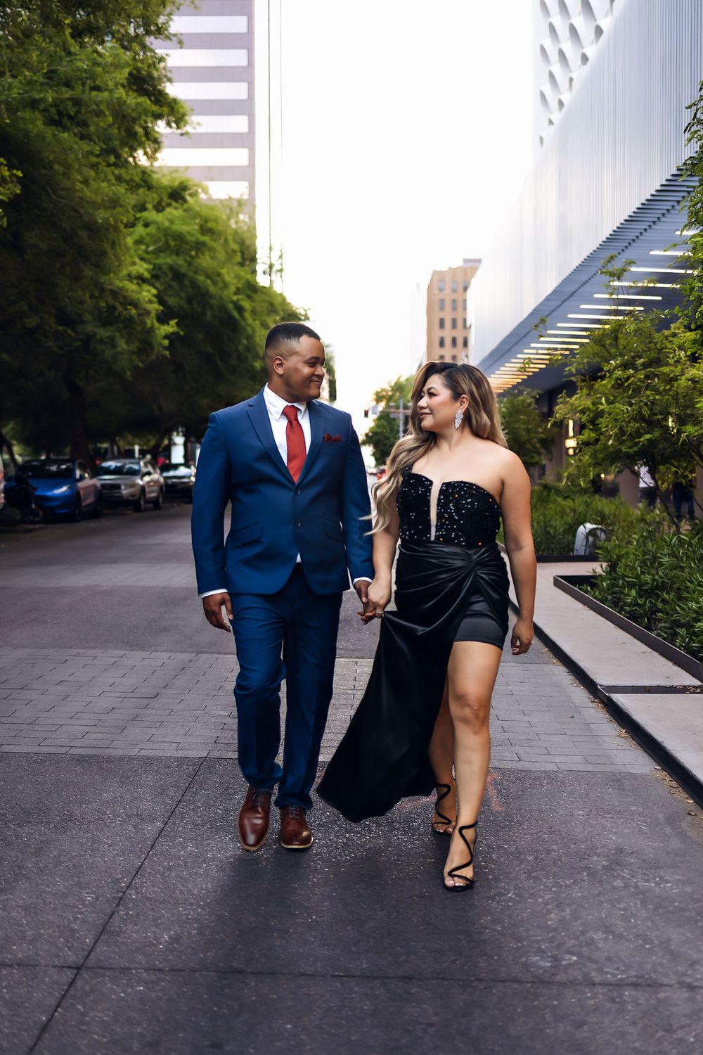 A couple dressed in formal attire walks together on an urban sidewalk in a downtown setting.