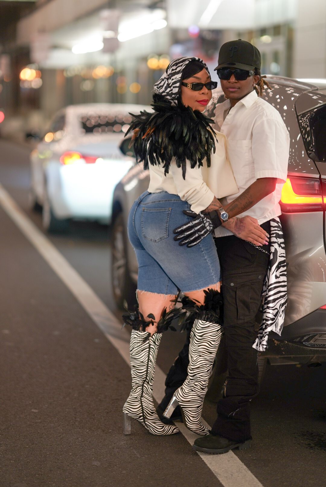 Fashionable duo in zebra print boots and feathers posing by vehicle.