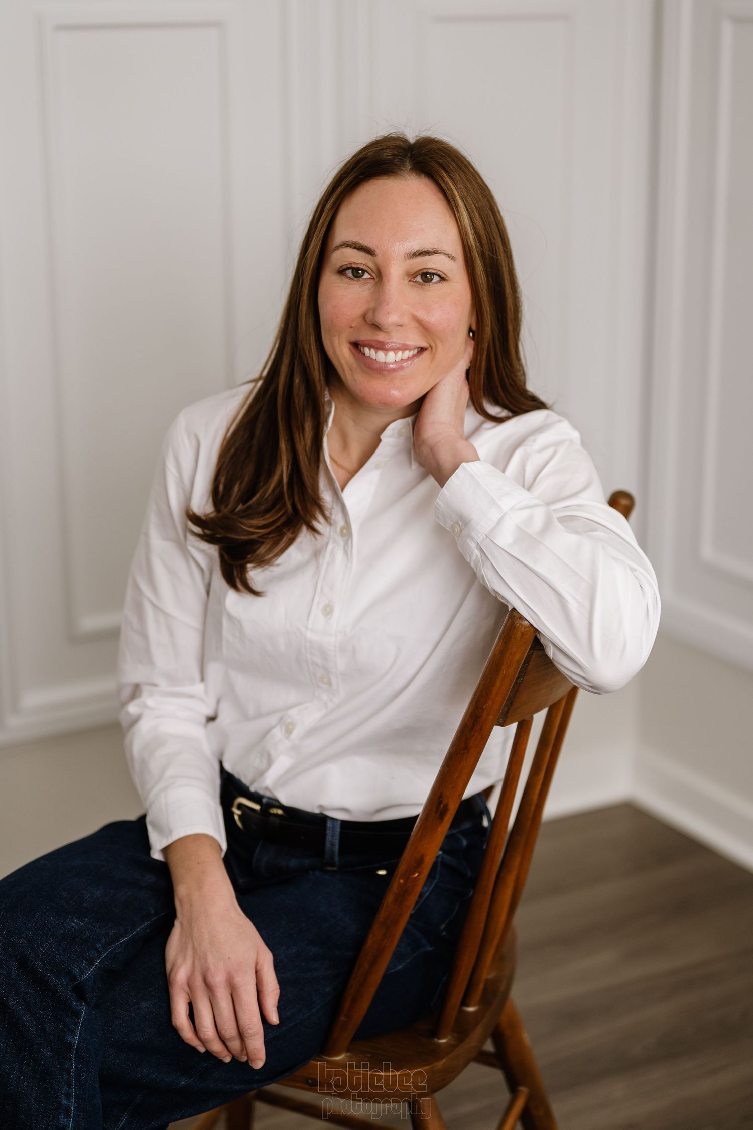 Professional portrait of someone seated on a wooden chair wearing a crisp white button-down shirt and dark jeans.
