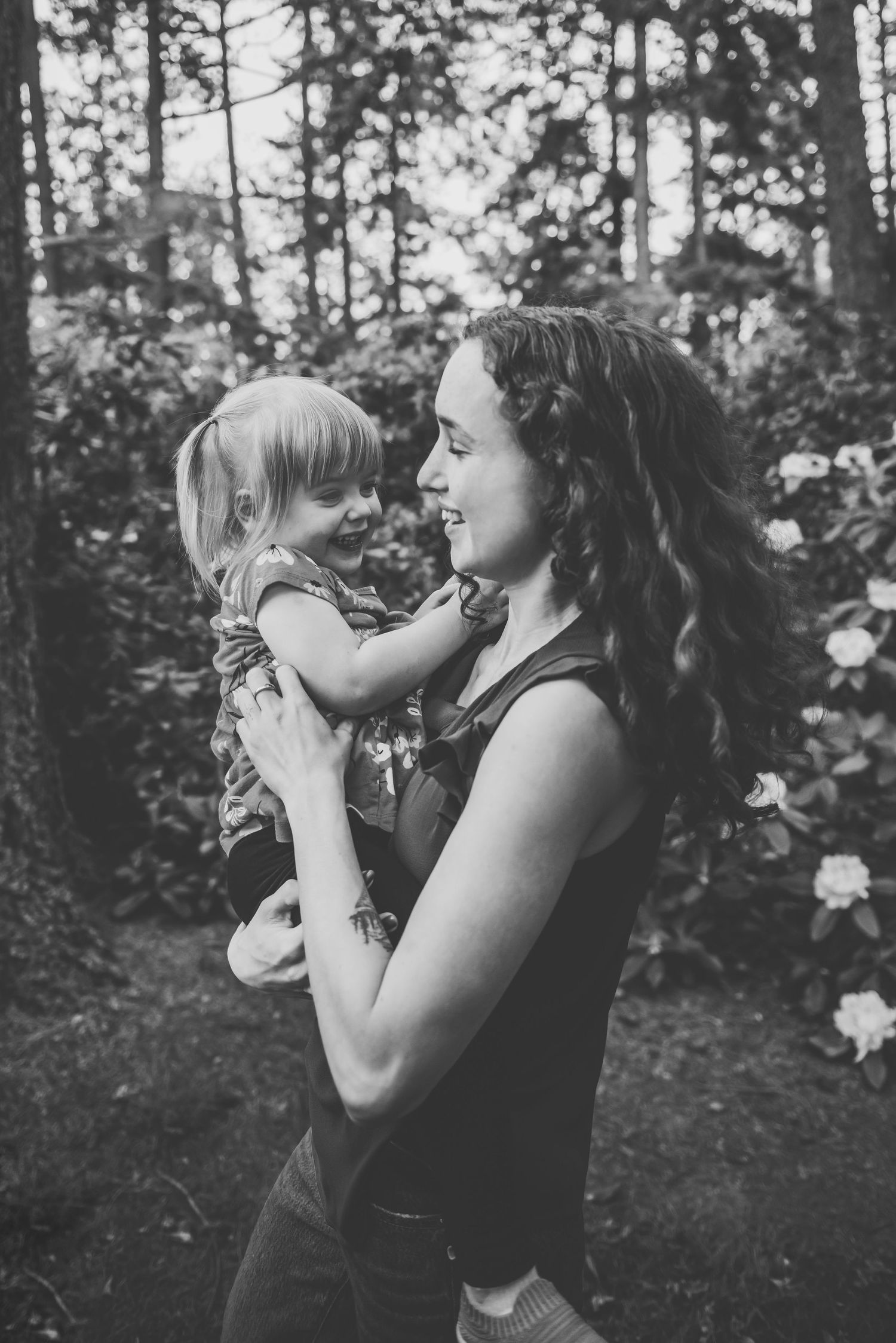 A tender black and white moment captured outdoors in a wooded garden setting with flowering bushes between mother and daughter.