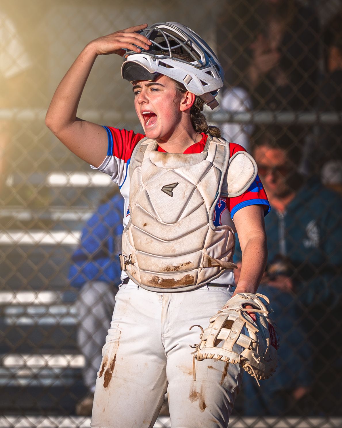 Youth baseball catcher in white uniform and protective gear shading eyes while looking up during sunny game.