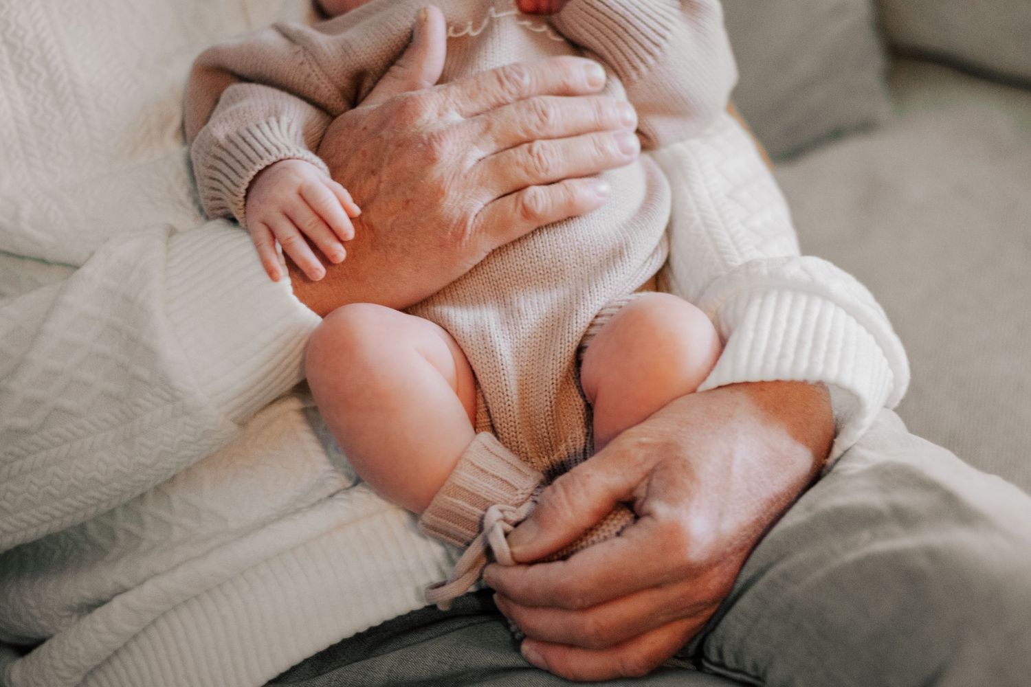 Small bare baby feet being held by gentle hands wearing a warm knit sweater on a cozy bed.
