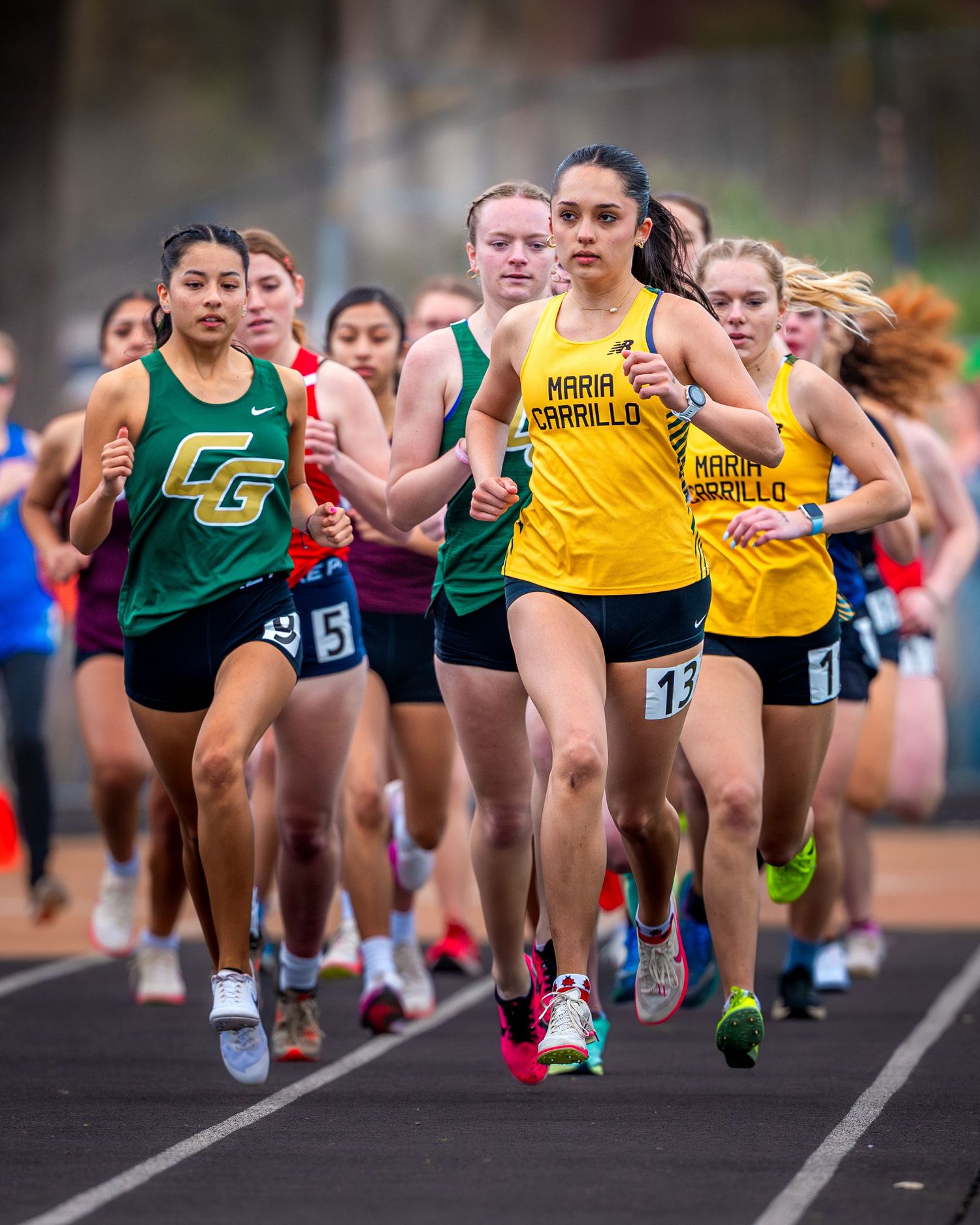 High school track athletes compete in a distance running event wearing yellow and green uniforms on an outdoor track.