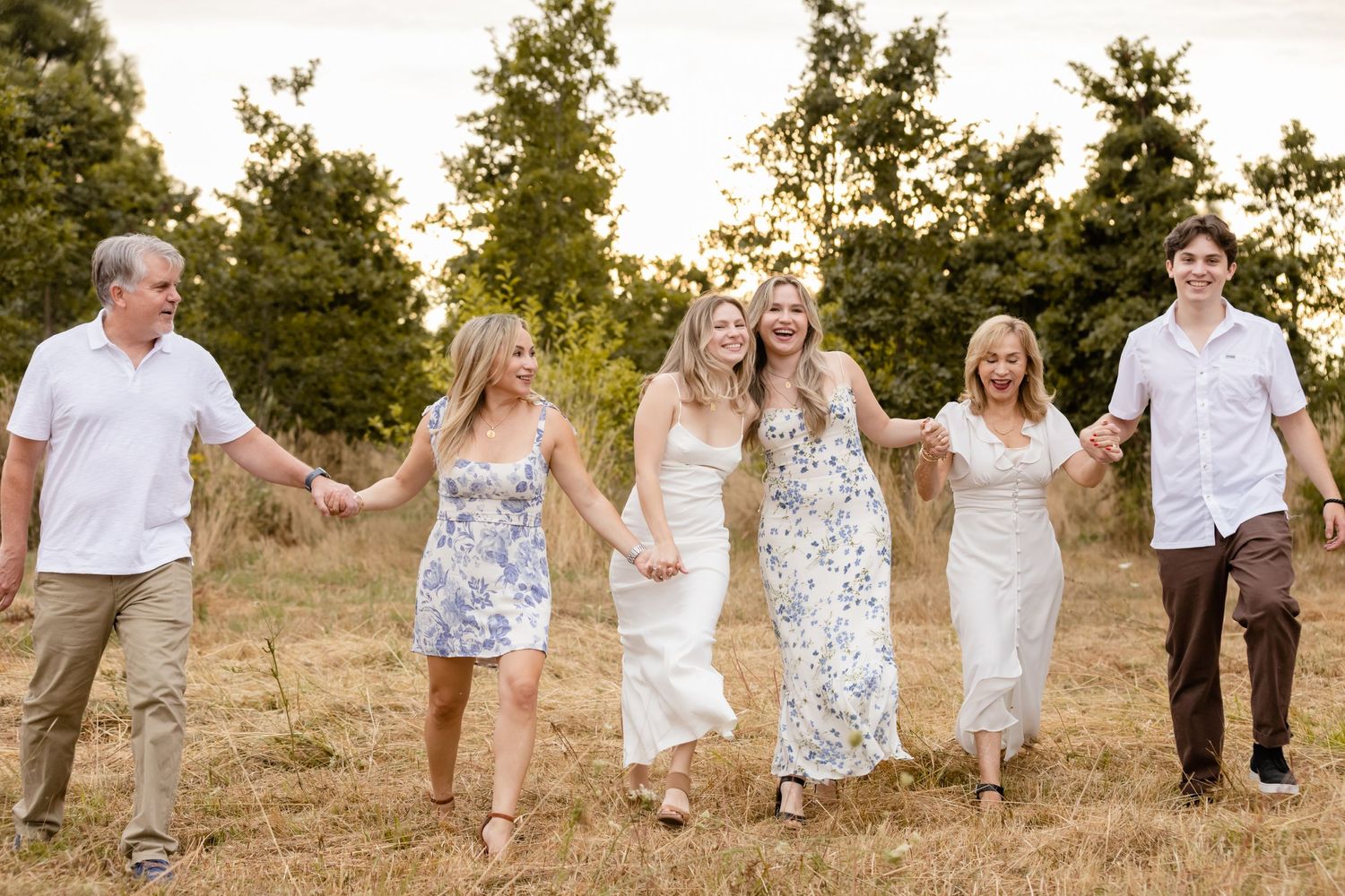A group holds hands walking together through a sunny field with evergreen trees in the background at sunset.