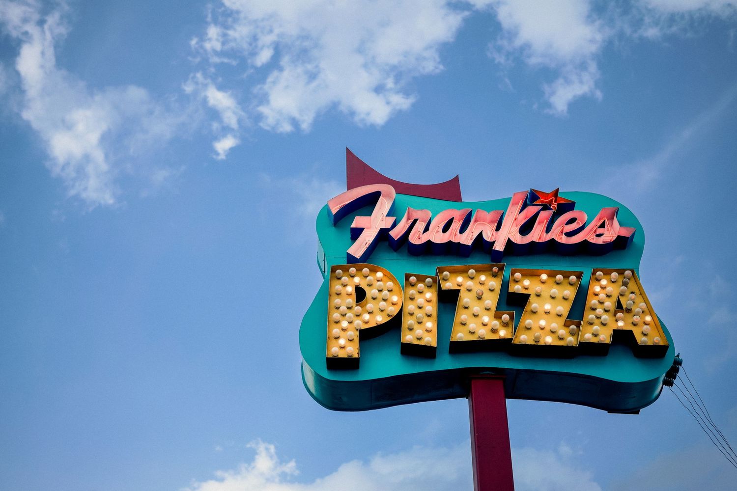 Vintage neon Frankie's Pizza sign illuminated against a bright blue sky with white fluffy clouds.
