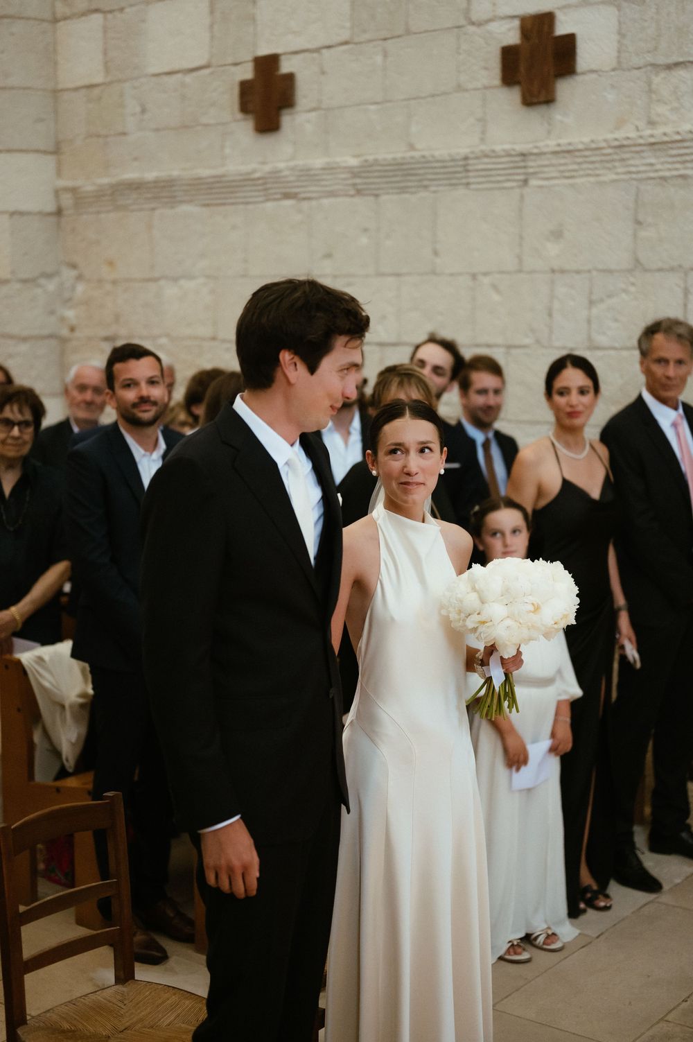 A wedding ceremony in a stone church with a couple in traditional white dress and black tuxedo surrounded by guests.