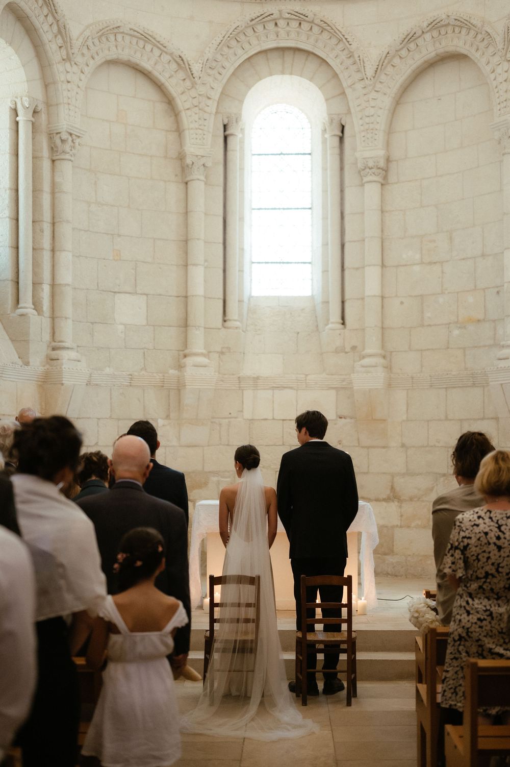 Wedding guests seated in rows inside a bright stone church with arched windows and columns.