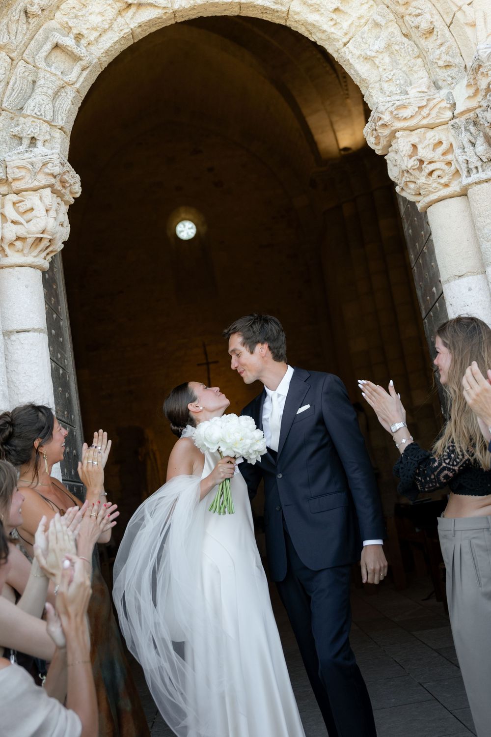 A wedding celebration in an elegant stone archway with guests in formal attire clapping and cheering.