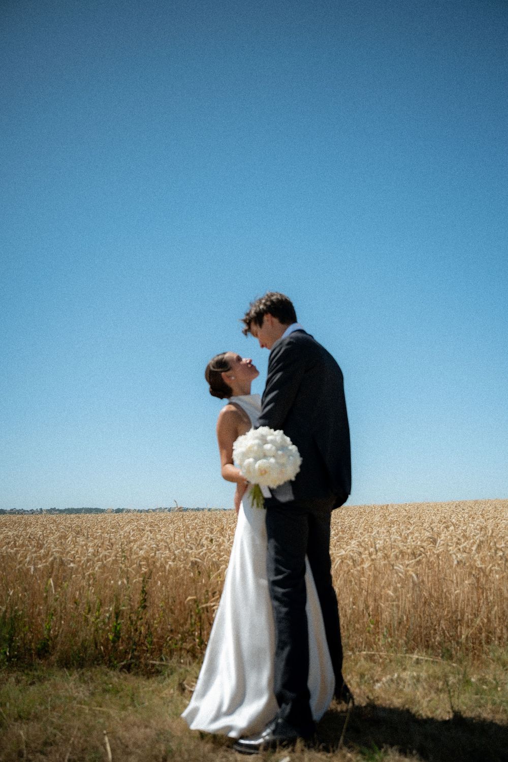 Newlyweds share an intimate moment in front of a golden wheat field under bright blue sky.