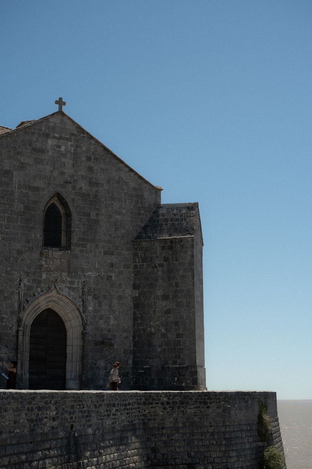 Ancient stone church with gothic arched entrance and cross on top against bright blue sky.