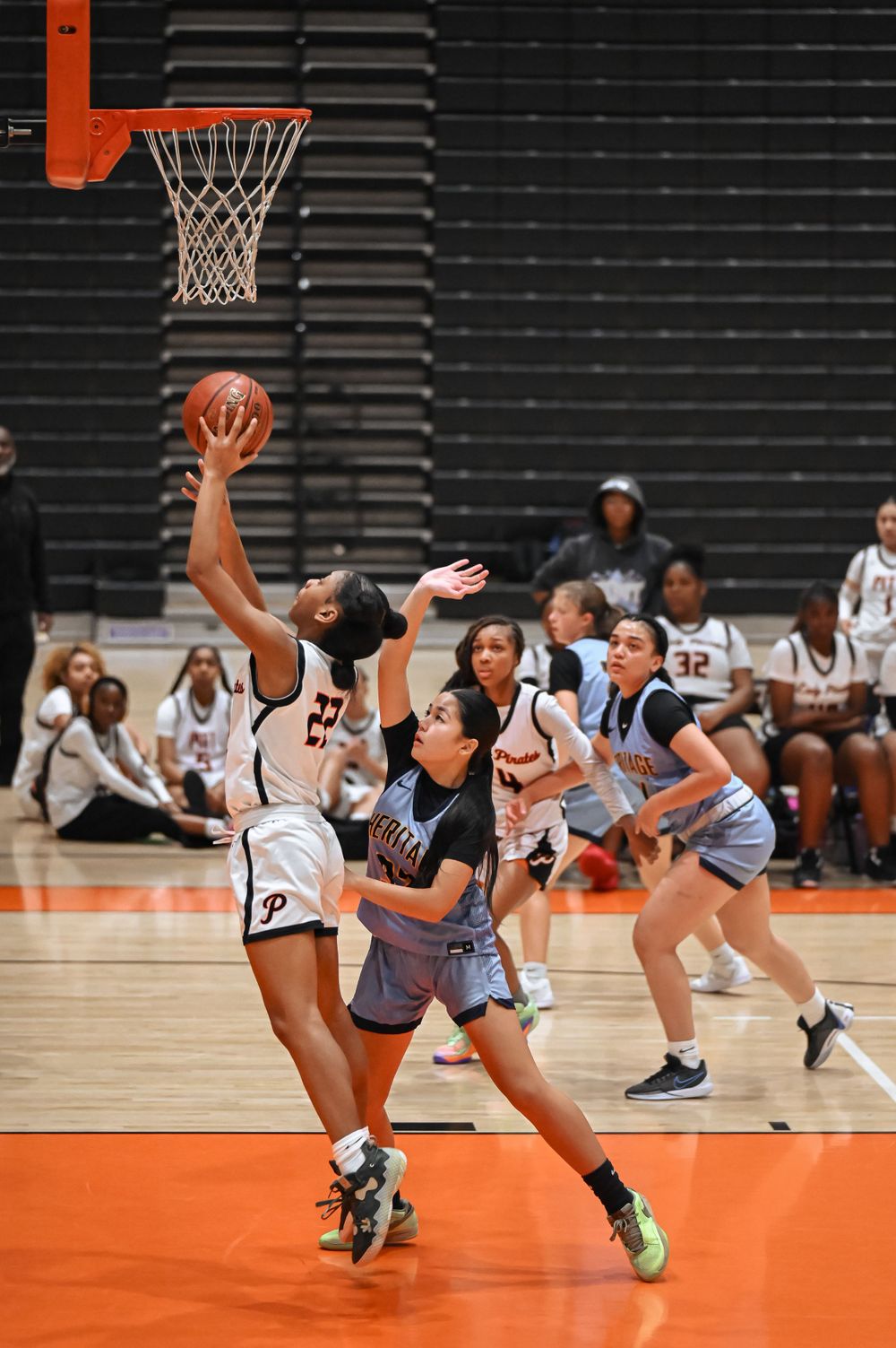 Players compete for a shot near the basket during an intense basketball game on an orange court.