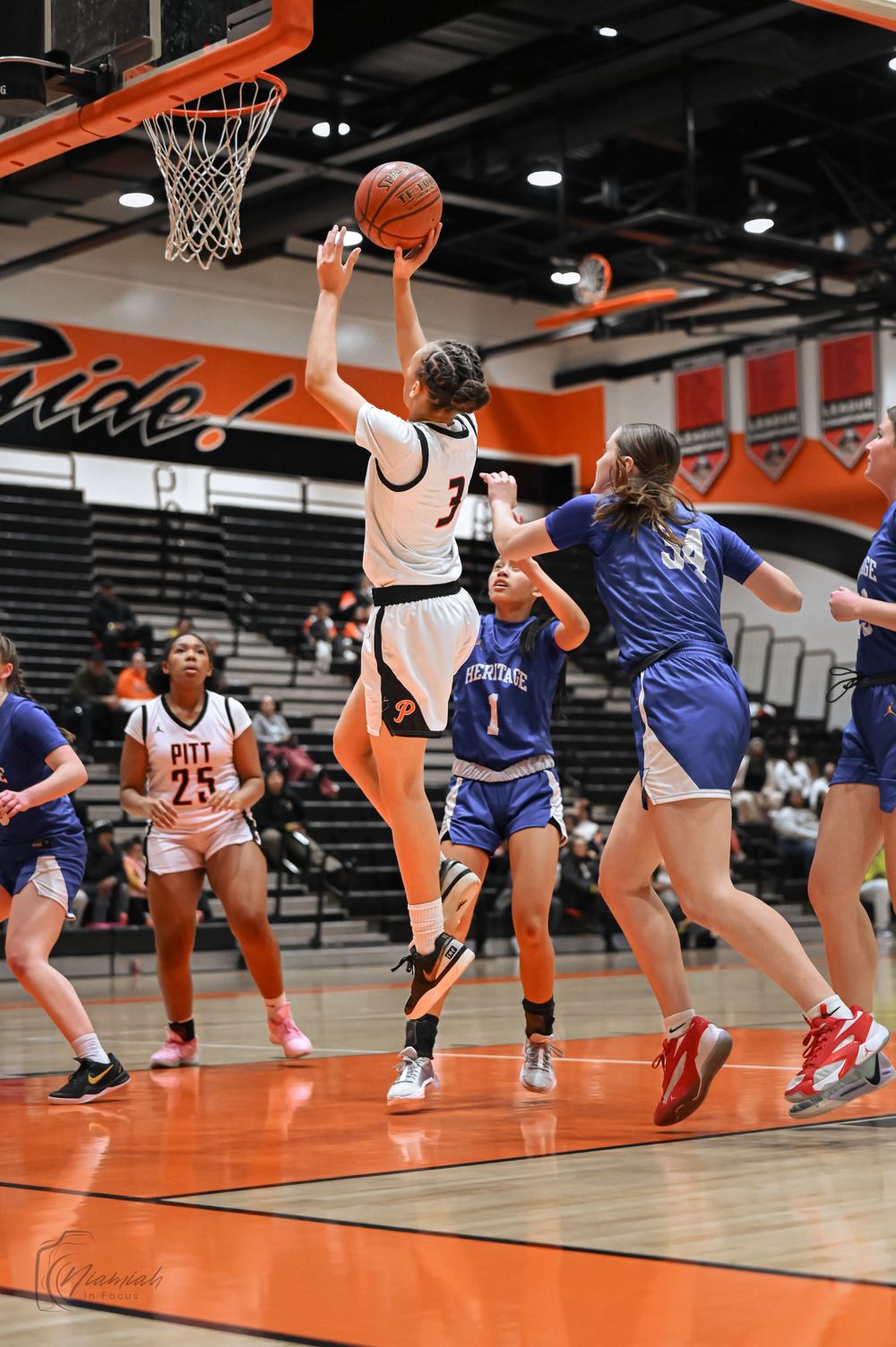 Basketball players jump for the ball during an intense game on an orange court with banners.