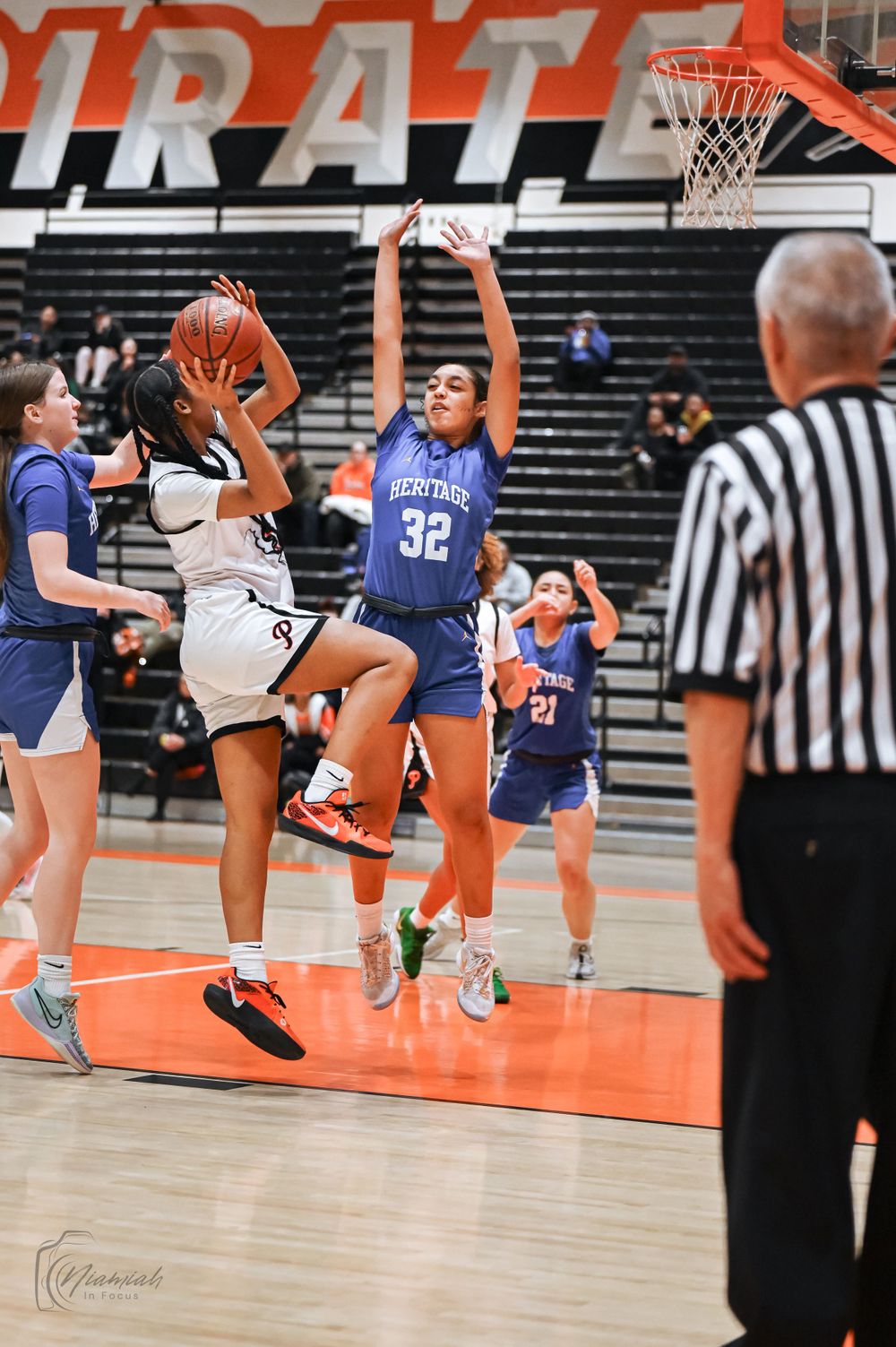Basketball players compete for a shot under the hoop as referee watches from sideline.