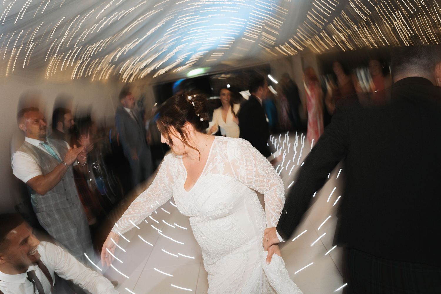 A blurred motion photo of a dance floor at a wedding reception shows swirling white dress movement in dim lighting.