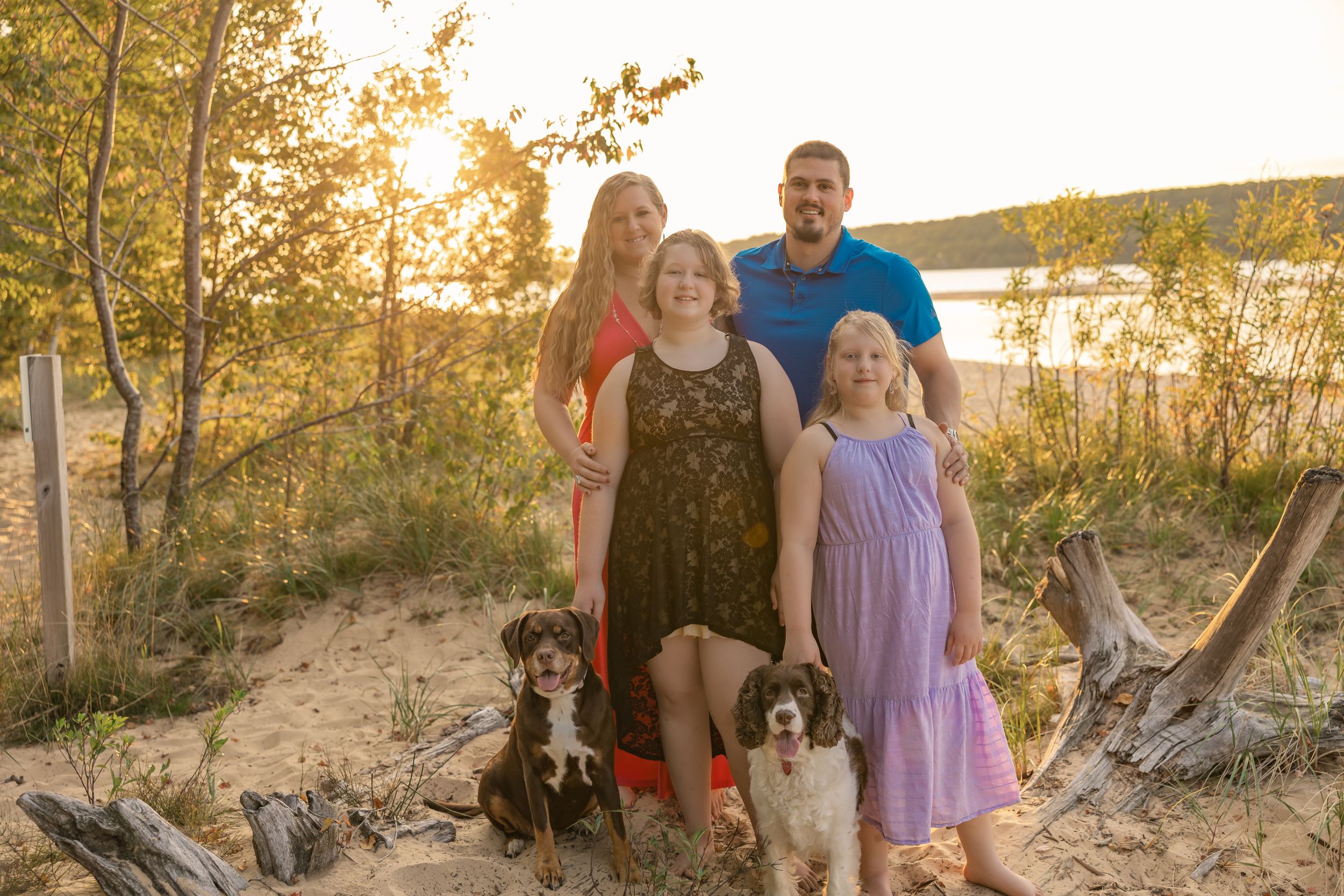 A group stands together with two dogs on a beach at sunset with water and trees in the background.