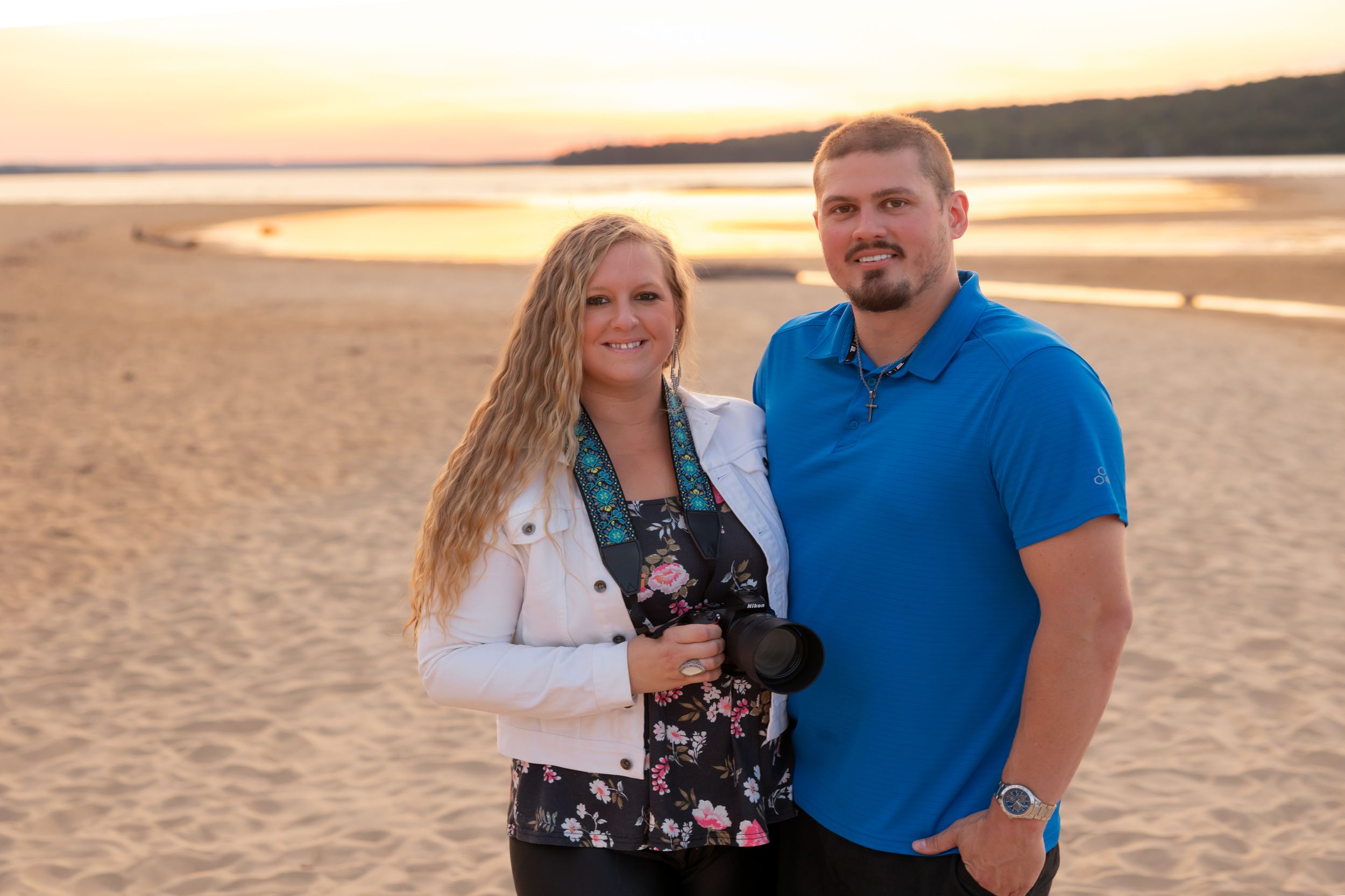 A couple stands together on a sandy beach at sunset with golden light glowing over the water.