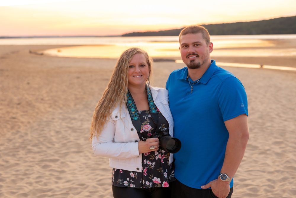 A couple stands together on a sandy beach at sunset with golden light glowing over the water.