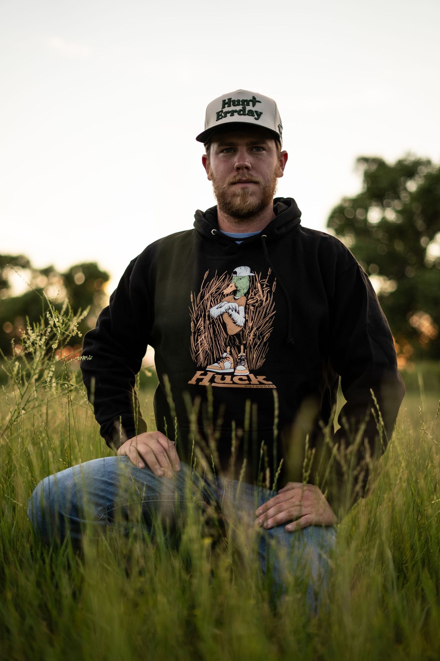 Someone wearing a black hoodie and baseball cap sits in tall grass during golden hour.