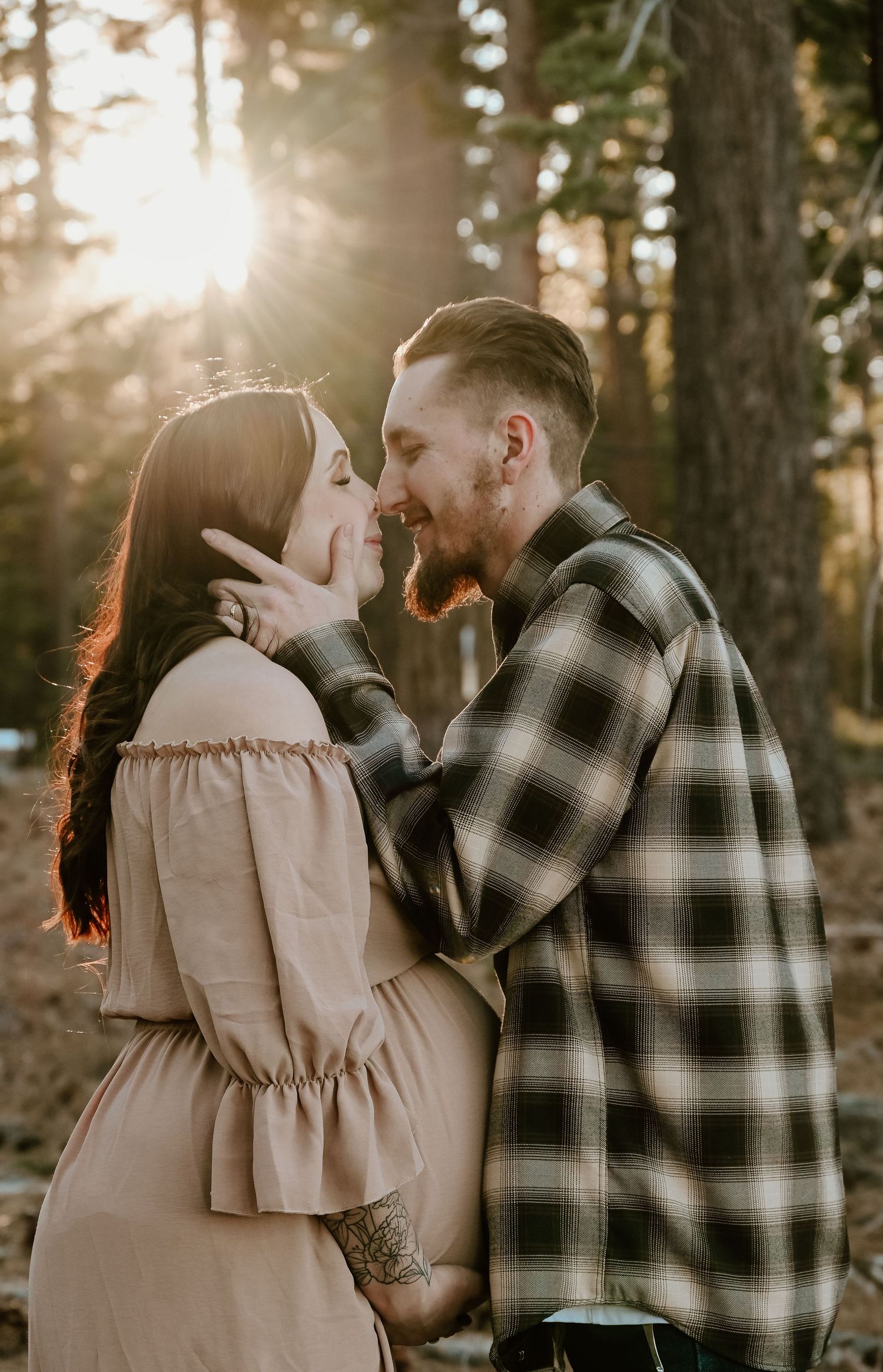 A couple shares a tender moment in a sunlit forest, with golden light streaming through the trees behind them.