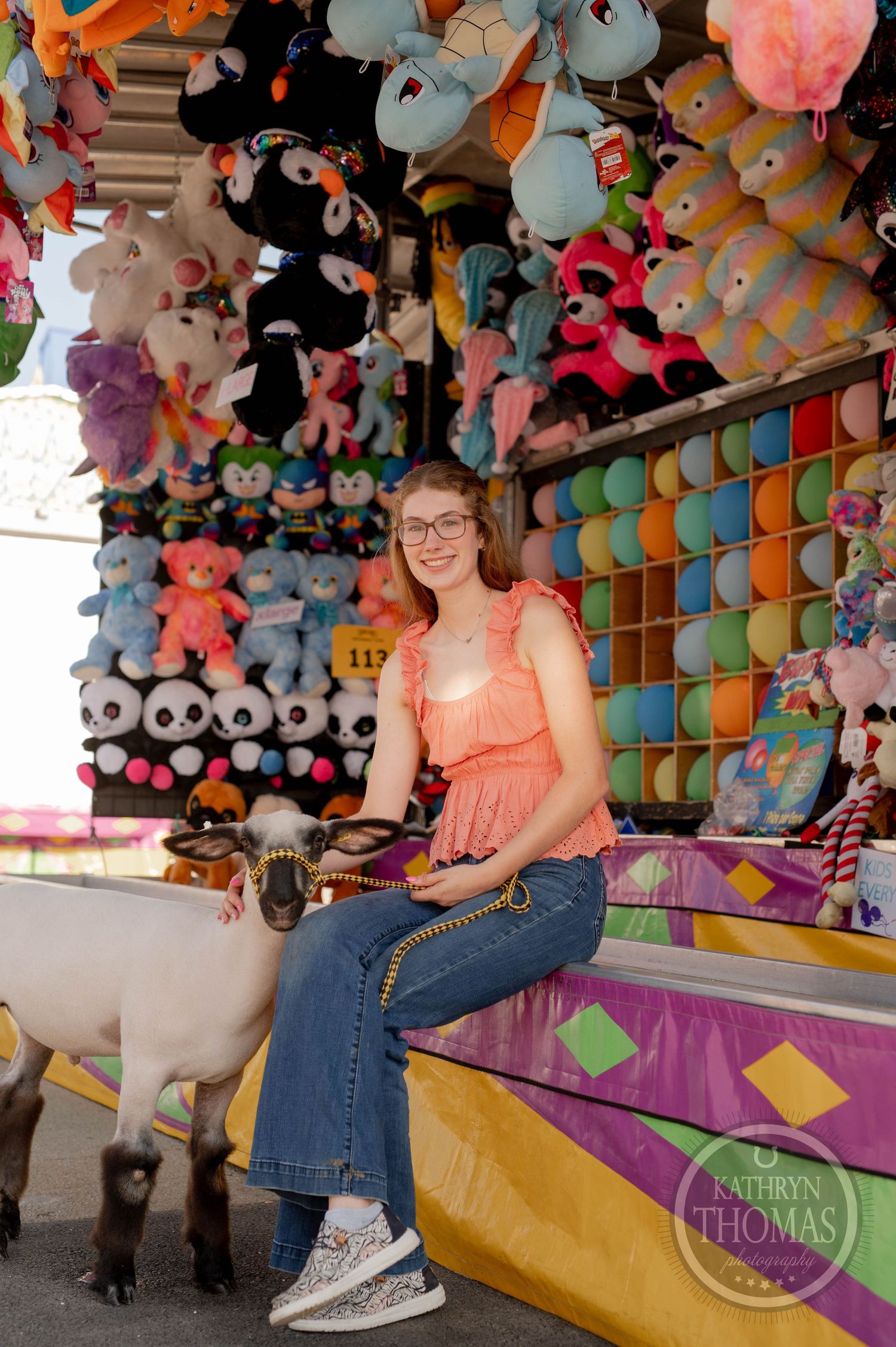 Woman sitting on carnival game booth bench surrounded by colorful plush toys.